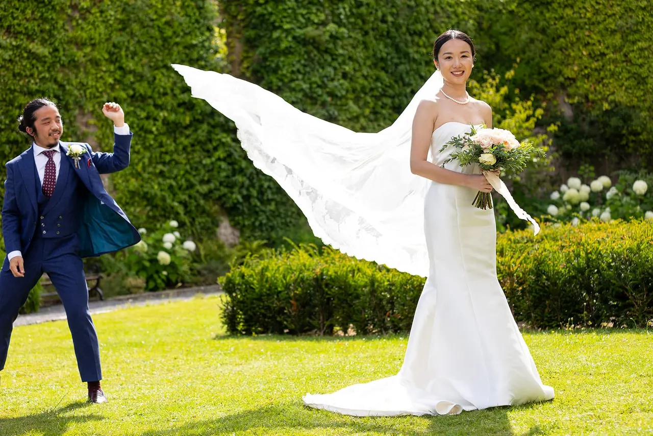 Bride and groom posing on a sunlit lawn with flowing white veil, holding bouquet, surrounded by manicured hedges and flowering borders.