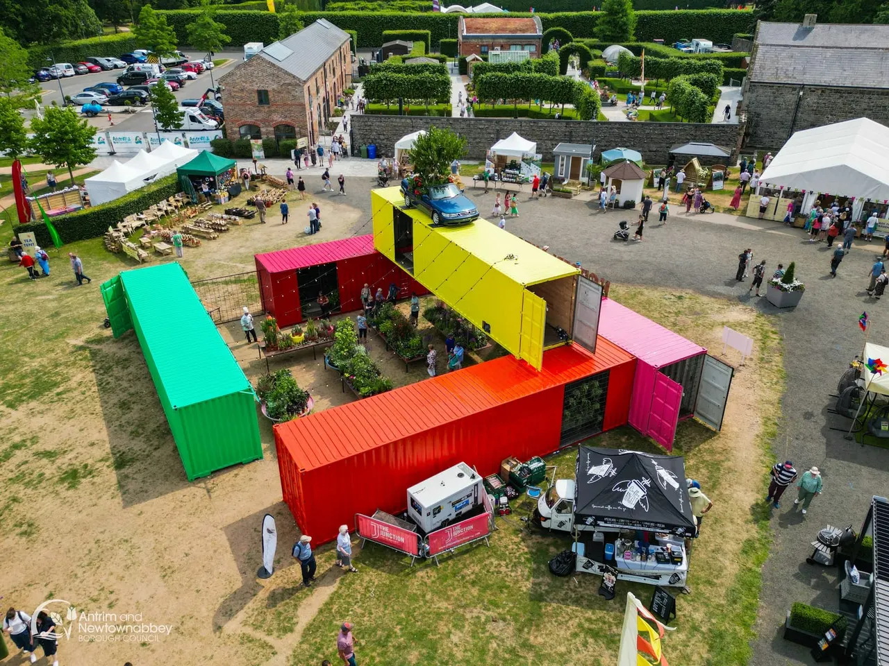 Aerial view of a leisure venue with colourful shipping container structures, visitors exploring exhibits, food stalls under white tents, and a car balanced on a yellow container roof.