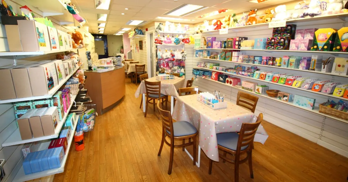 A bright, welcoming ceramic studio interior with shelves stocked with colourful paints and supplies, craft tables with chairs, and decorative items hanging from the ceiling.