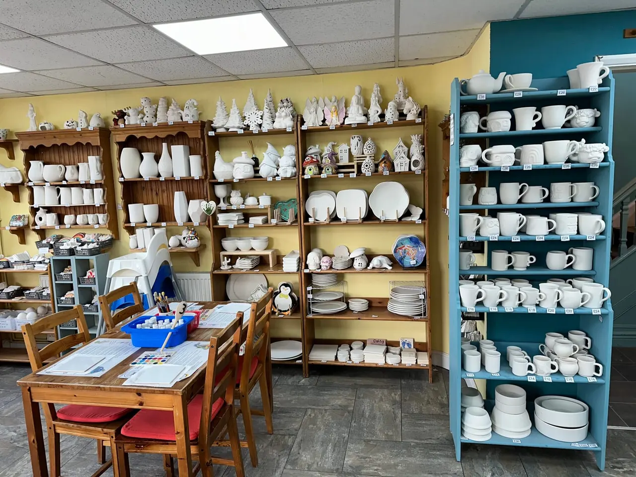 A pottery painting studio with wooden shelves displaying numerous white ceramic pieces, decorative houses, and figurines, plus a turquoise shelving unit stocked with white crockery and mugs.