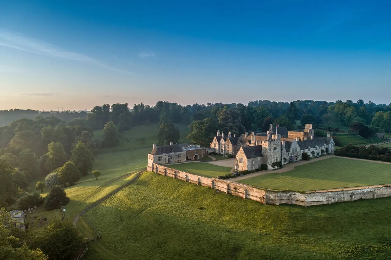Grand stone castle with multiple towers set within defensive walls, surrounded by sprawling green grounds and woodland under clear blue sky.