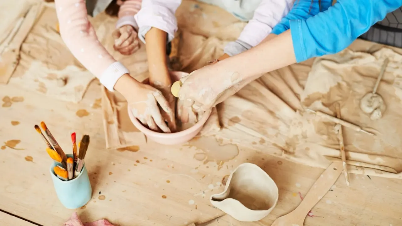 Children's hands working with clay at a pottery table, with paintbrushes and ceramic pieces nearby.