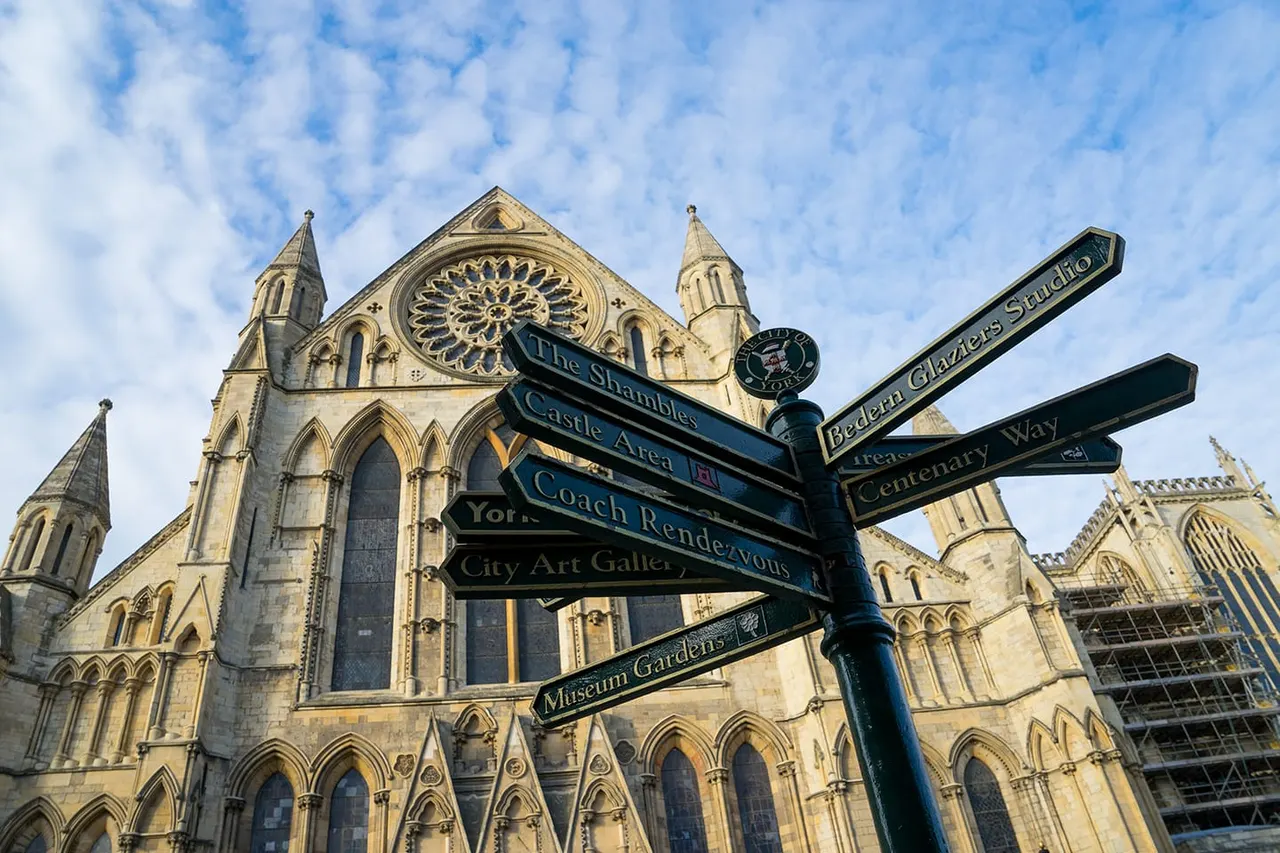 Directional signpost with multiple arms in front of a Gothic stone cathedral under blue sky with white clouds.