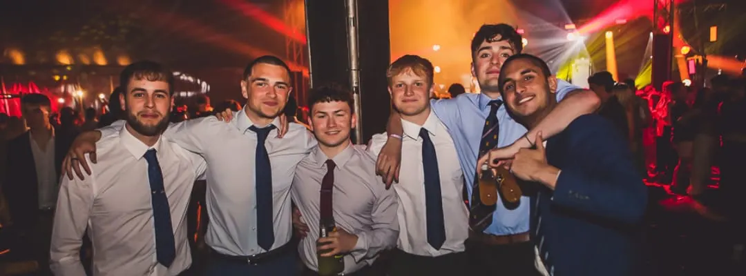 Group of men in smart casual attire posing together at a nightclub with colourful lighting and crowds in background.