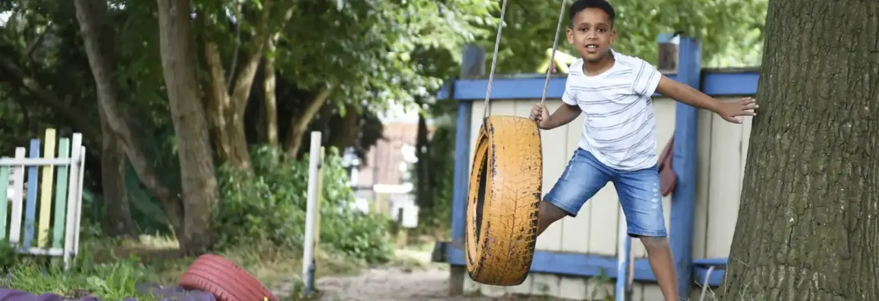 Child playing on a tyre swing suspended from a tree in a wooded outdoor play area.