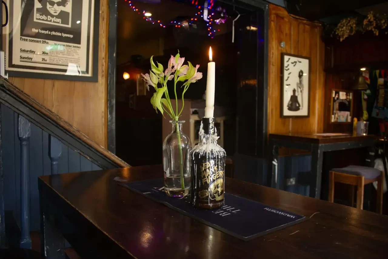 Bar counter with a lit candle in a decorated bottle holder next to a vase of pink flowers, with concert posters and coloured lights visible in the warm-lit interior.