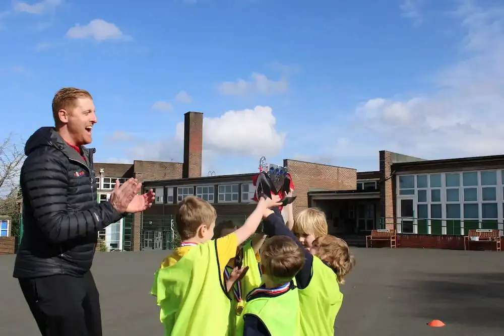 An adult instructor in a black jacket engages with a group of children in high-visibility vests outdoors on a school playground, holding up a sports item.