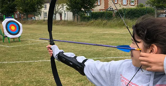 Young person drawing back a bow and arrow aimed at a target on a grass field with buildings and trees in the background.