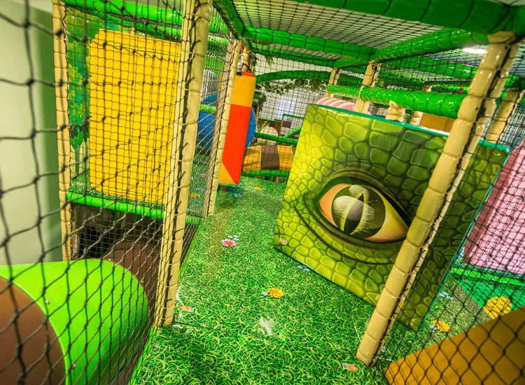 Indoor soft play maze with green netting, colourful obstacles, and a large dinosaur eye graphic on the wall.