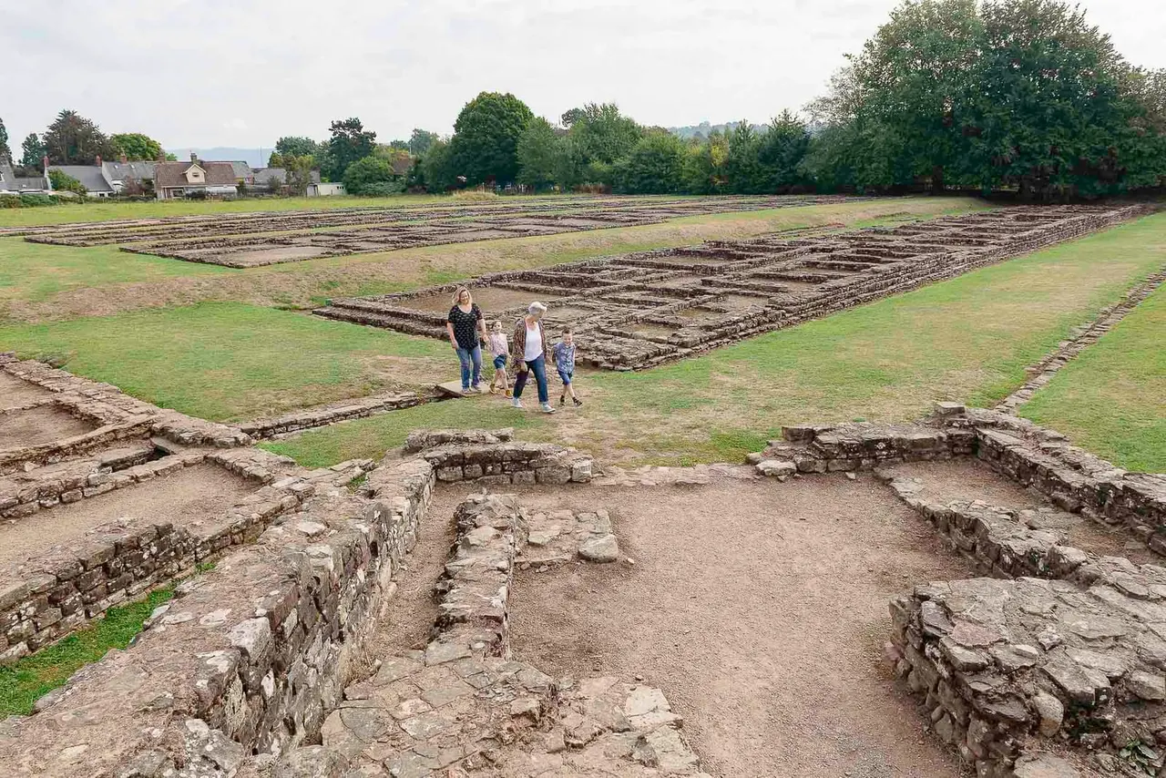 Family walking amongst Roman stone ruins and earthwork remains at an archaeological site near countryside cottages.