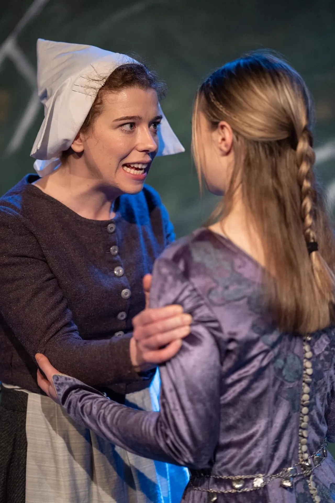 Two young performers in period costumes engaged in a theatrical scene, one wearing a white bonnet and the other with braided hair.