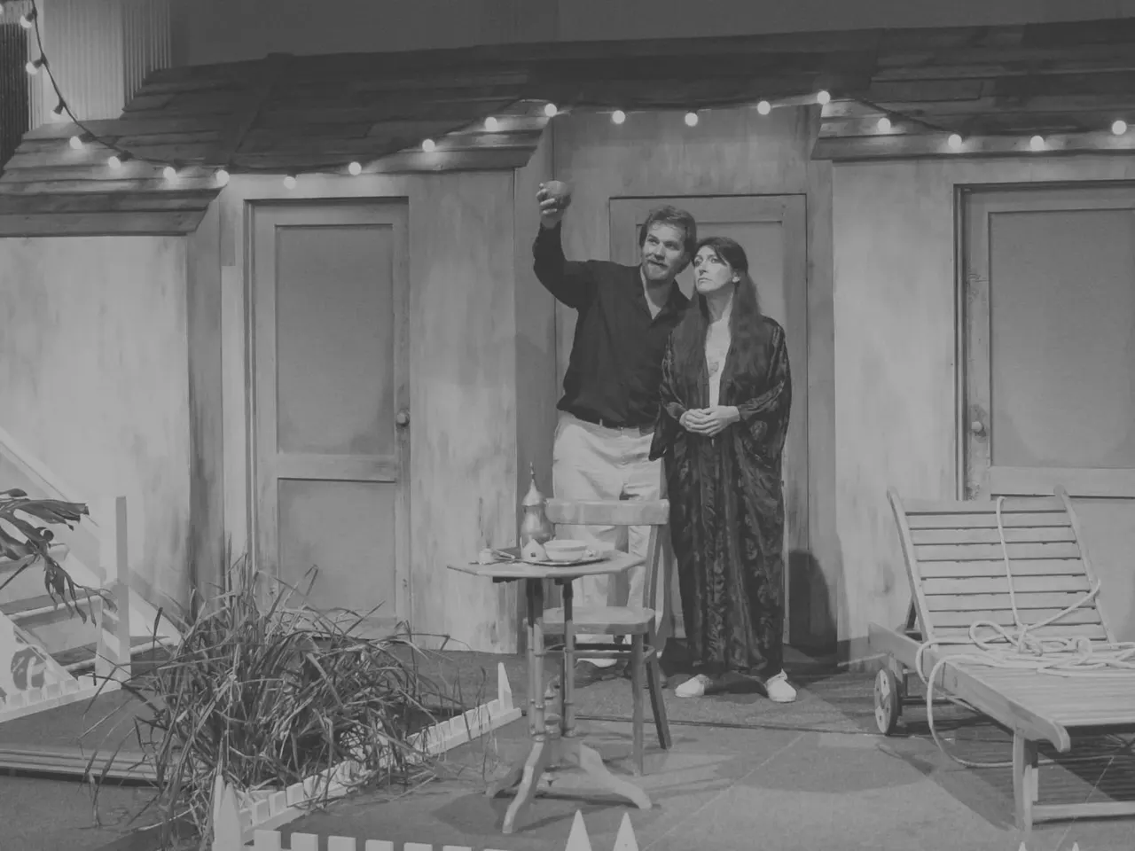 This is a PHOTOGRAPH. Let me describe what it shows:

Two people stand on a patio set decorated with string lights, surrounded by loungers and potted plants, in black and white.