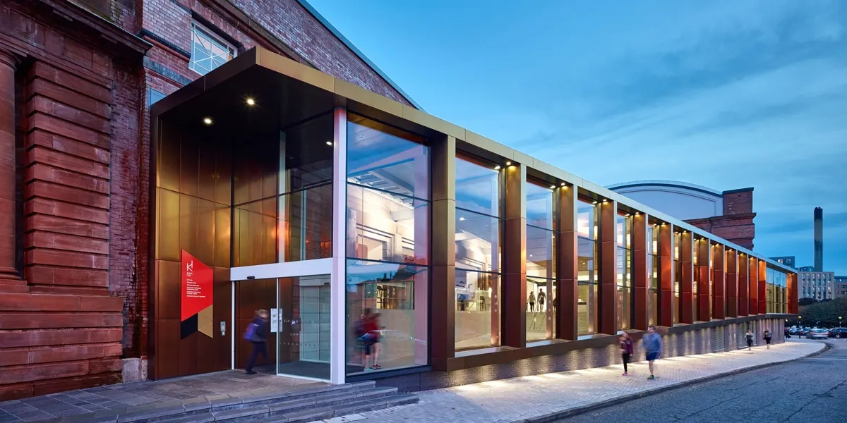 Modern glass-fronted building with red brick exterior at dusk, visitors walking past illuminated entrance.