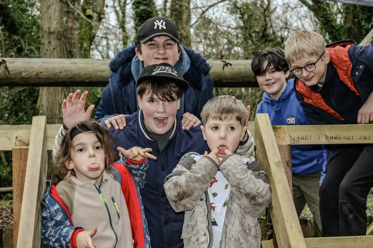 A group of six children posing playfully inside a wooden structure with trees in the background.