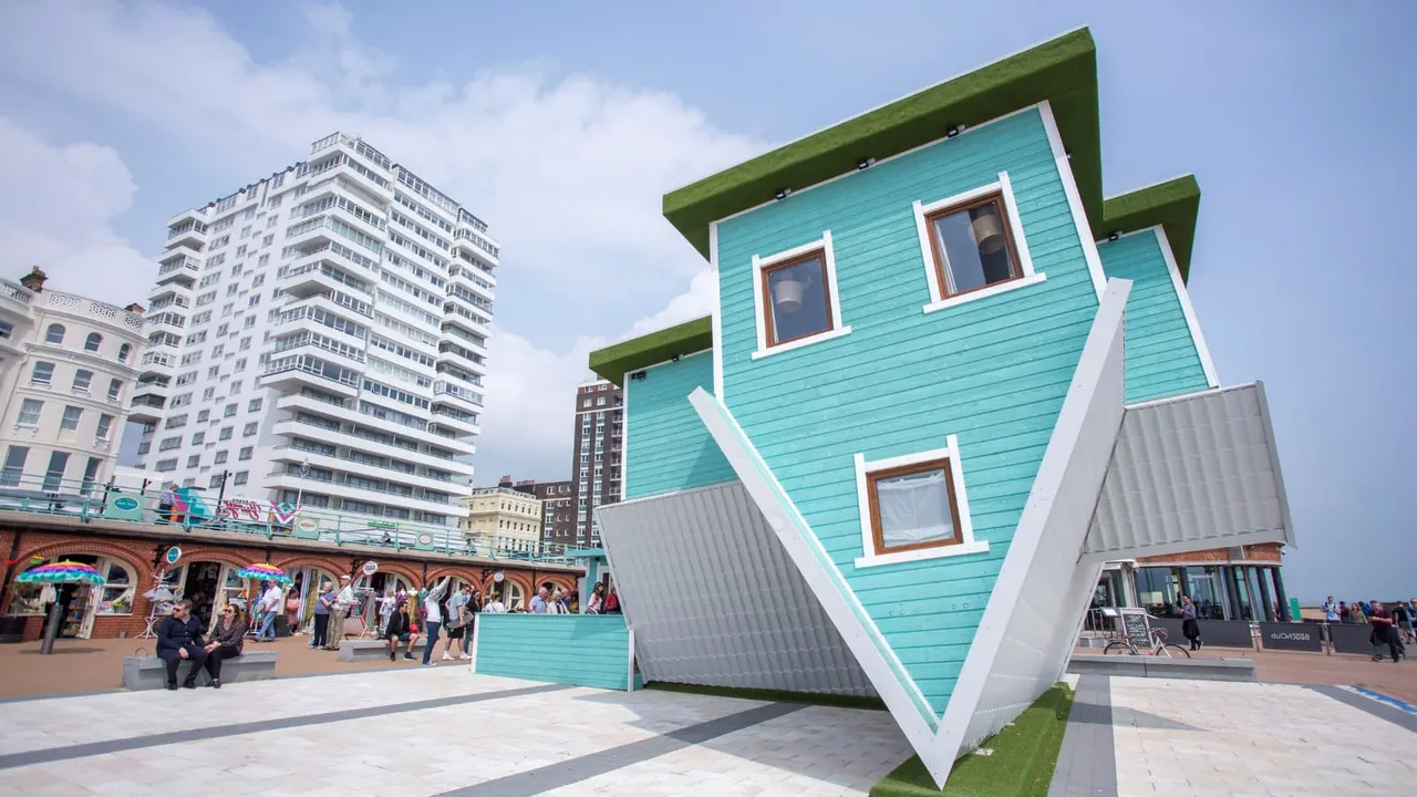 An upside-down turquoise house with green roof overlooking Brighton seafront promenade and beach buildings.