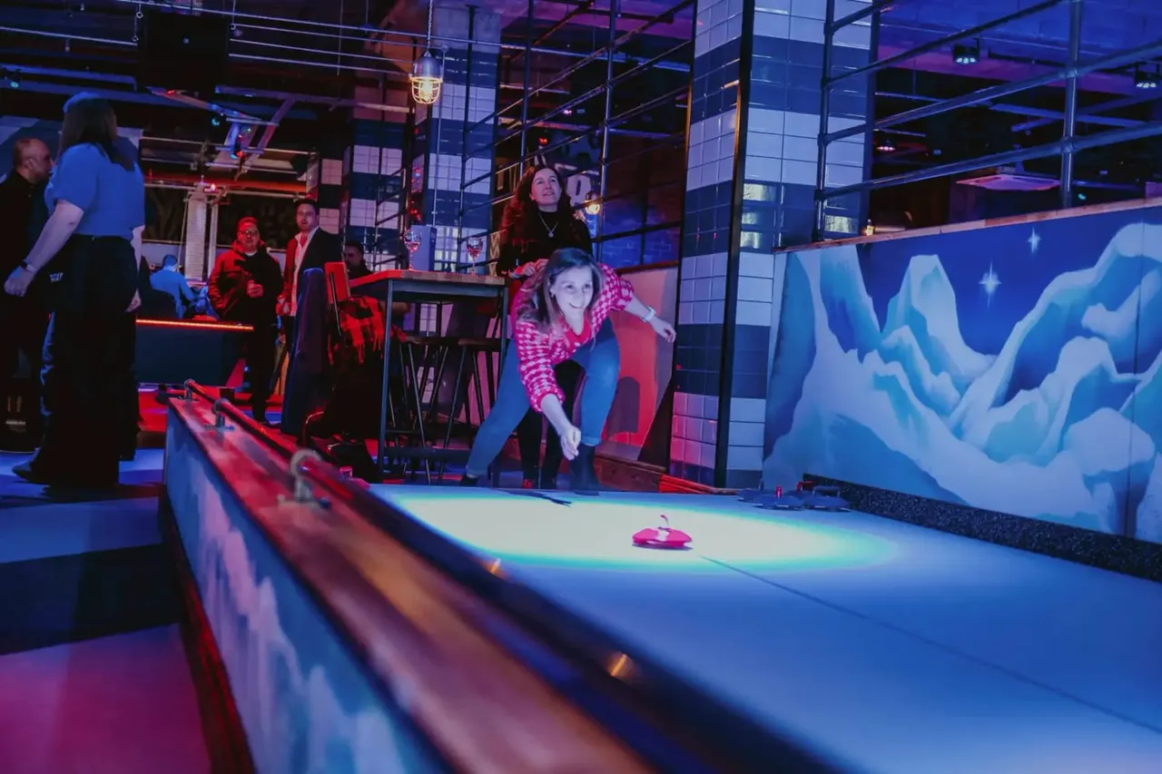 People playing curling on an illuminated indoor rink with neon lighting and digital screens.
