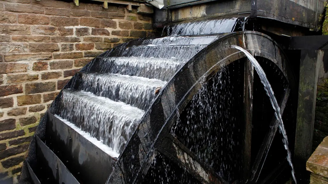 Water cascades over the steps of an old mill wheel turning against a stone building wall.