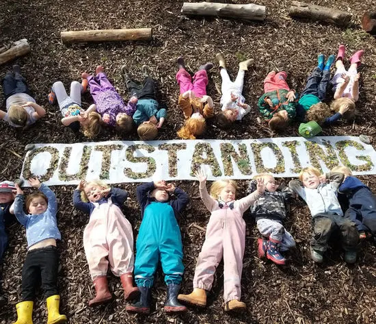 Children lying on ground holding a banner reading "OUTSTANDING" at a forest school.