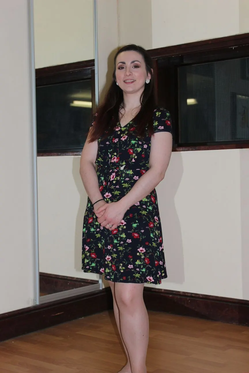 Woman in floral dress standing indoors against cream and dark wood panelled wall.