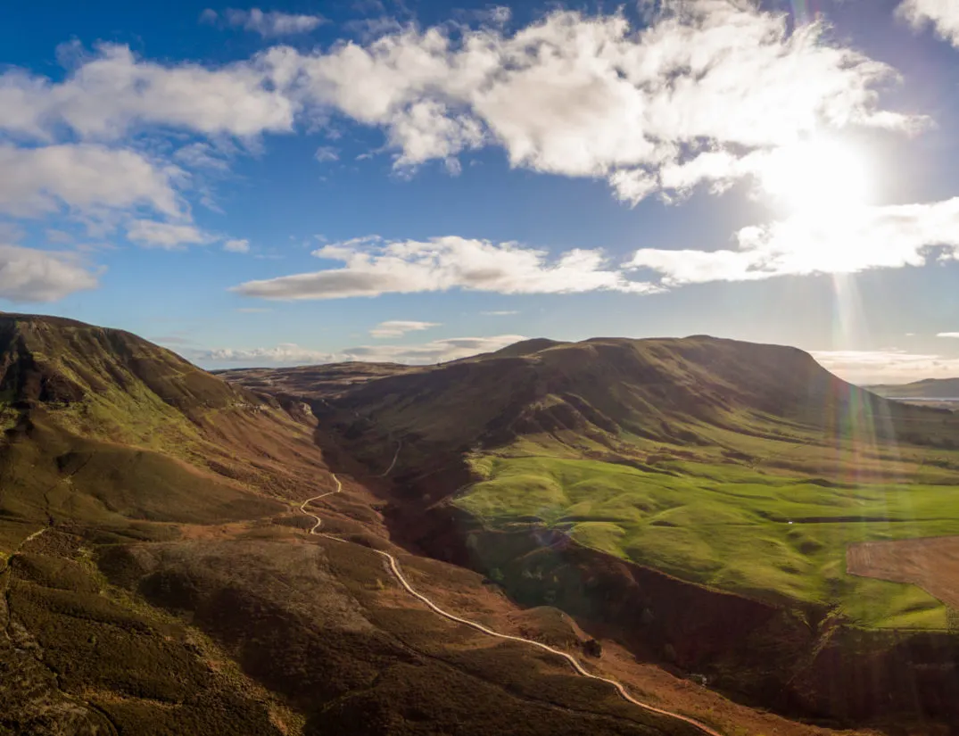 Expansive Highland valley landscape with rolling mountains, green moorland and a winding river under blue sky with white clouds.