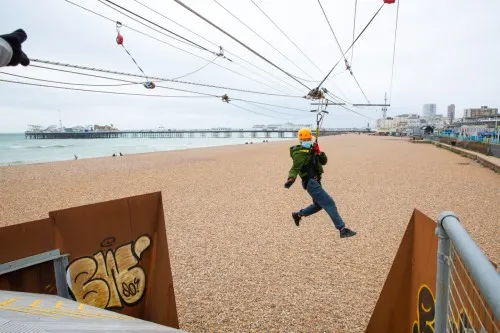Person ziplining across a beach with a pier and seafront buildings in the background.