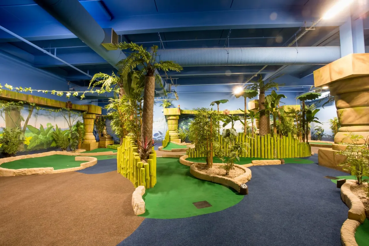 Indoor tropical-themed mini golf course with green fairways, palm trees, yellow structures and jungle backdrops.