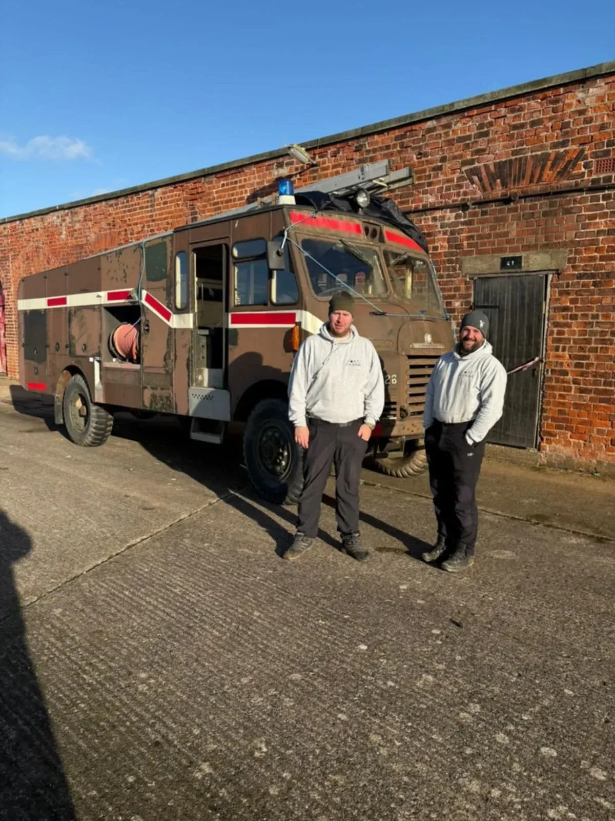 Two people stand in front of a vintage military ambulance against a brick building.