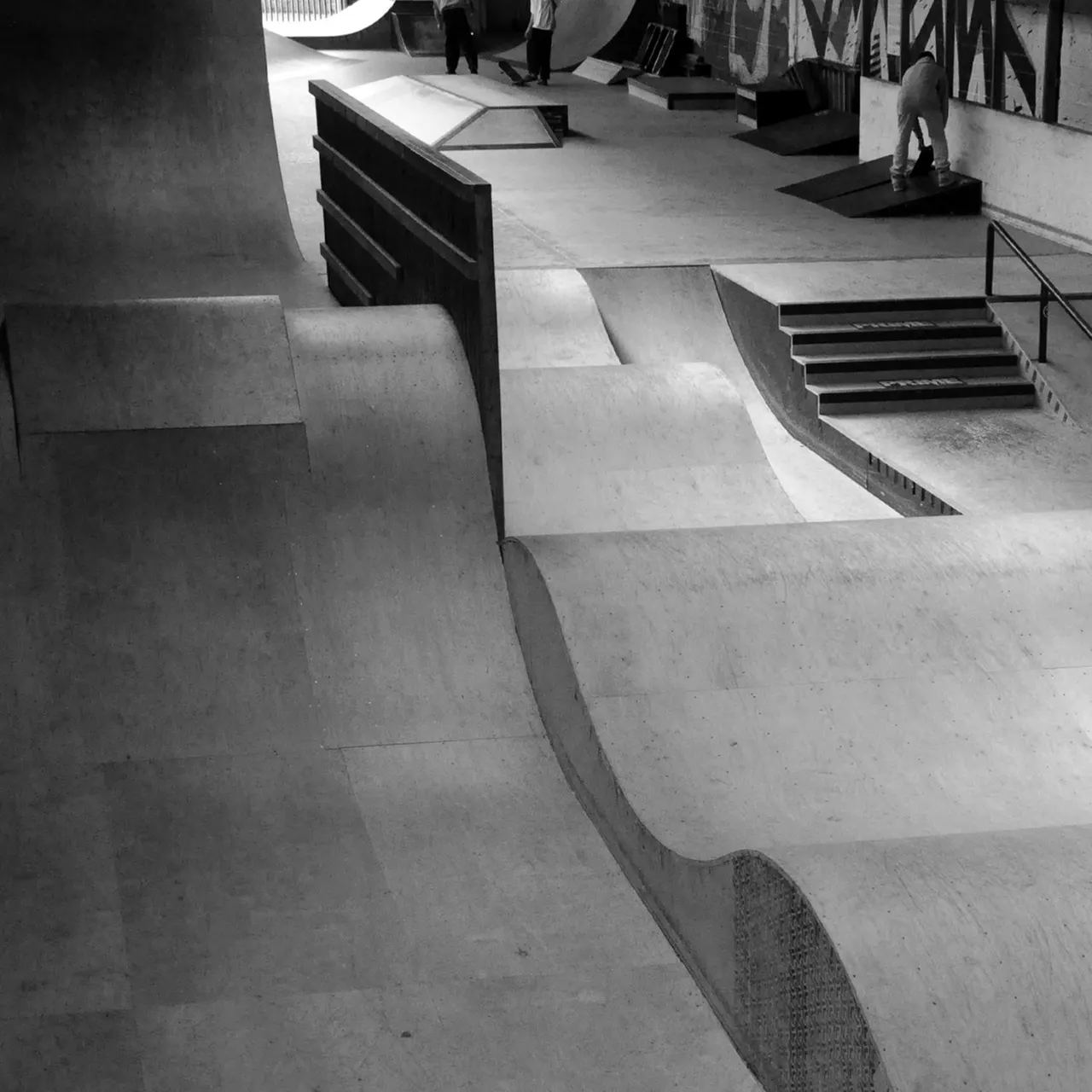 Skate park with concrete bowls, ramps, and stairs, a few people scattered around the empty facility.