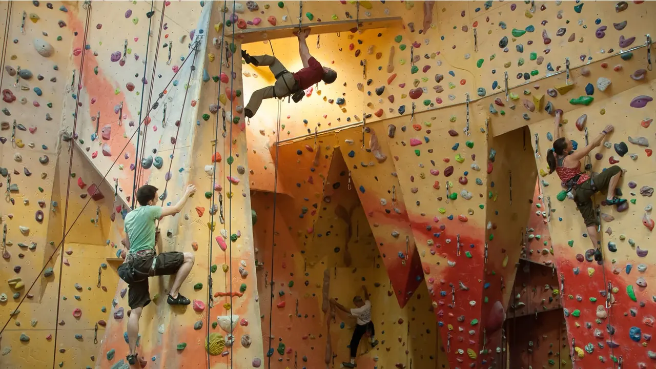 Multiple climbers on indoor rock climbing walls covered in colourful handholds, some using ropes for top-rope climbing.