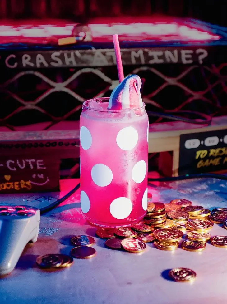 A pink polka-dot cocktail with a straw sits among arcade game tokens on a neon-lit arcade bar counter.