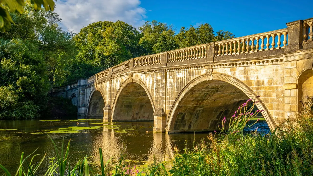 Historic stone bridge with three arches spanning a river, surrounded by woodland and flowering plants on a sunny day.
