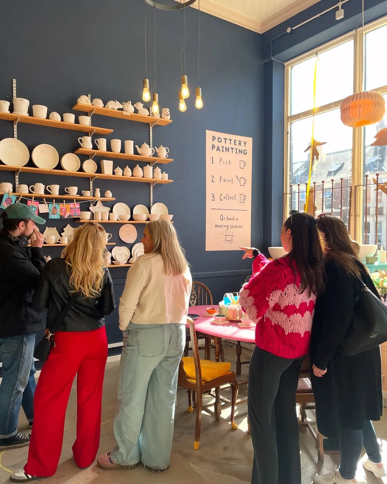 A pottery painting studio with visitors gathered to view ceramic pieces displayed on wooden shelves against a dark blue wall, with instructions posted nearby.