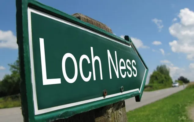 Green directional road sign pointing to Loch Ness with clear blue sky and countryside road visible behind.