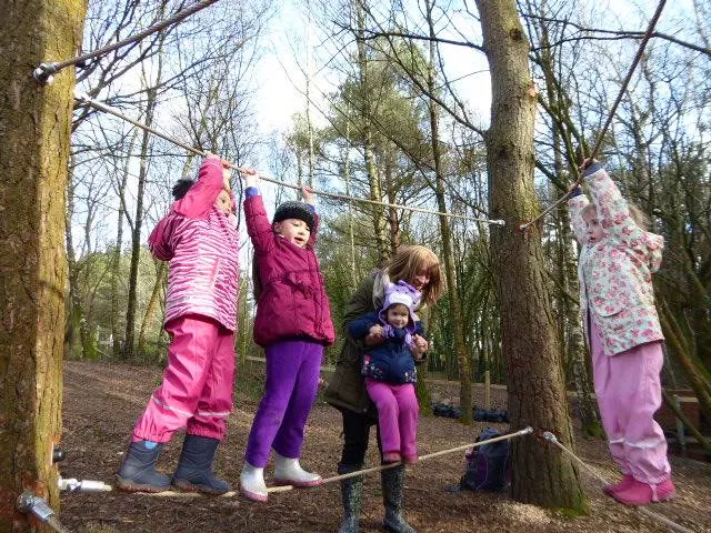 Children in brightly coloured clothing playing on a rope and pulley course among trees in a woodland setting.