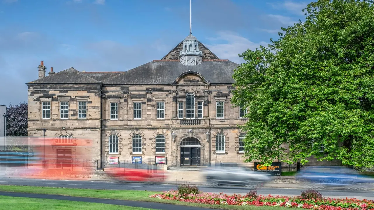 Historic stone theatre building with cupola, fronted by flower beds and passing traffic on sunny day.