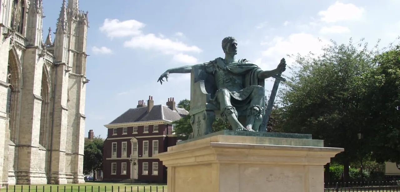 Bronze statue of a robed male figure on a stone plinth, with Gothic cathedral and red brick building behind.