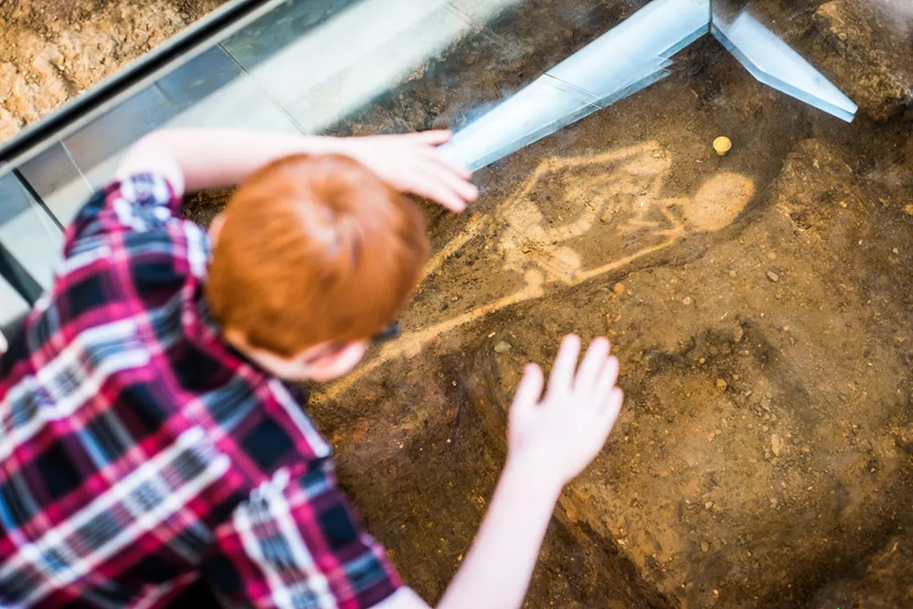 Child in checked shirt touching footprints in sand within an indoor display tank.