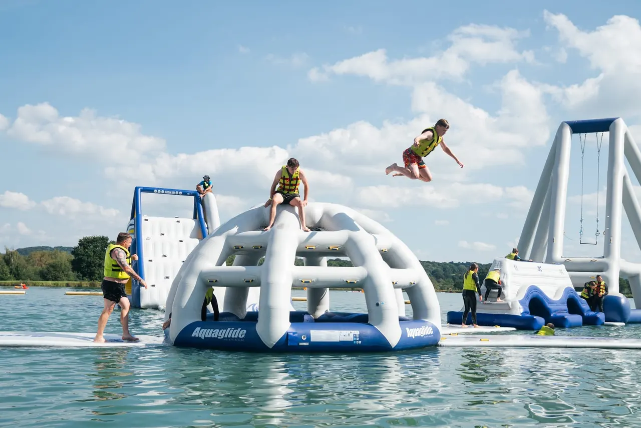 People in life jackets enjoy inflatable water park attractions on a lake under blue sky with white clouds.