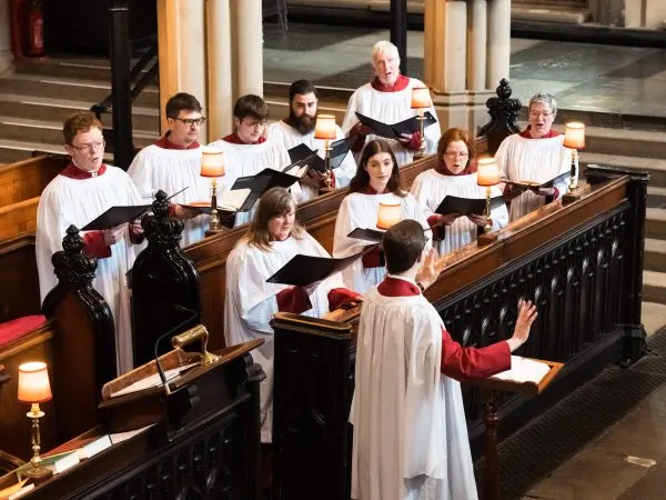 A choir in white robes and red stoles singing together in a church chancel, led by a conductor.