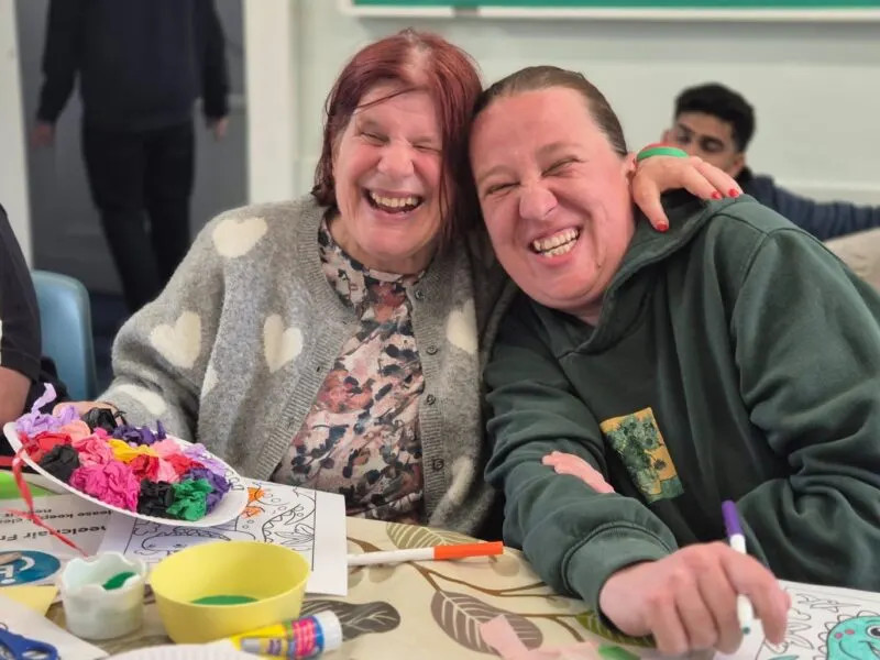 Two women smiling at a table during a craft activity with colouring materials and decorative items.