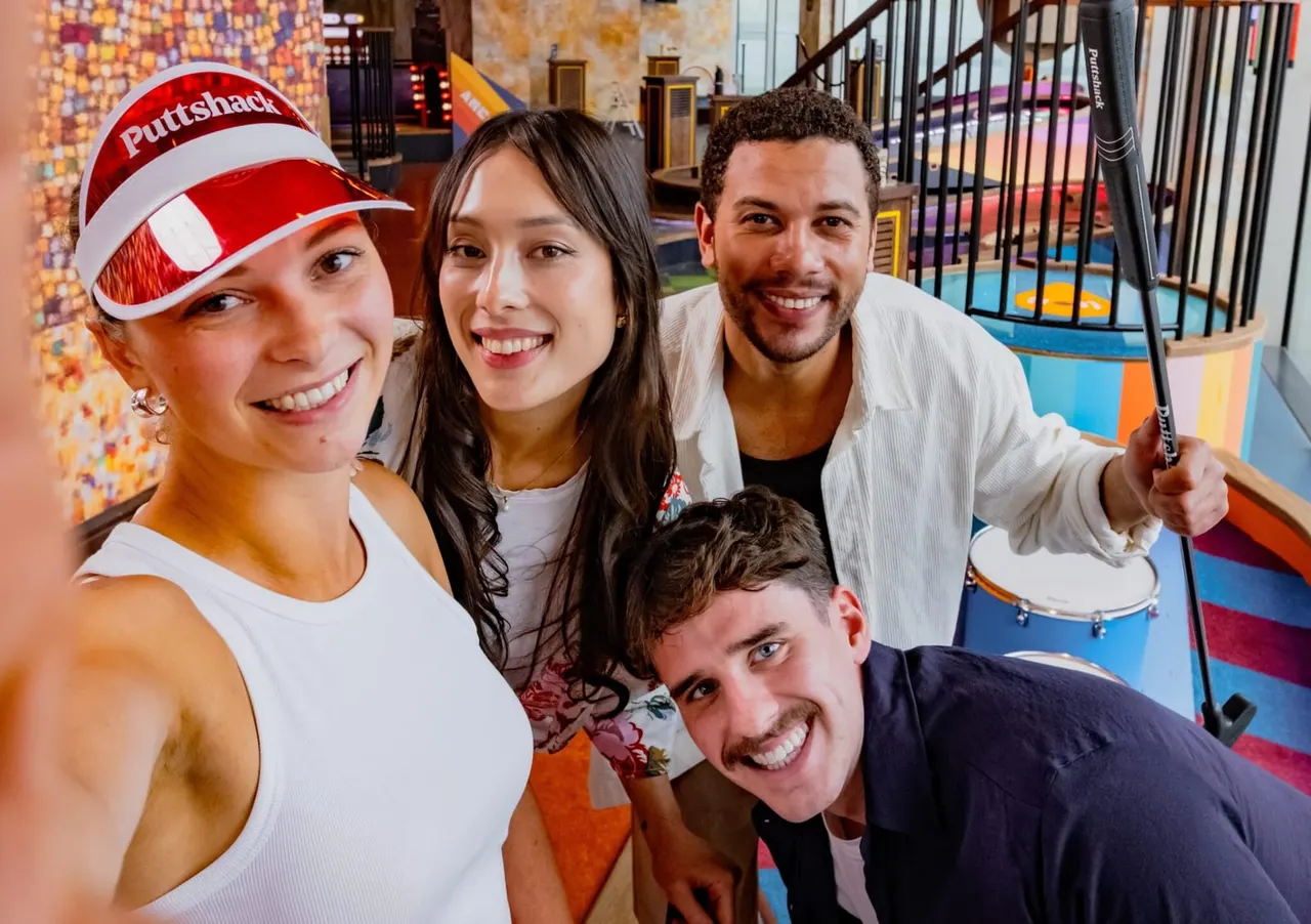 Four smiling people posing together inside a colourful indoor mini golf venue with colourful flooring and obstacles visible in the background.