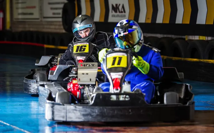 Two go-kart racers wearing helmets and numbered bibs compete on an indoor track.