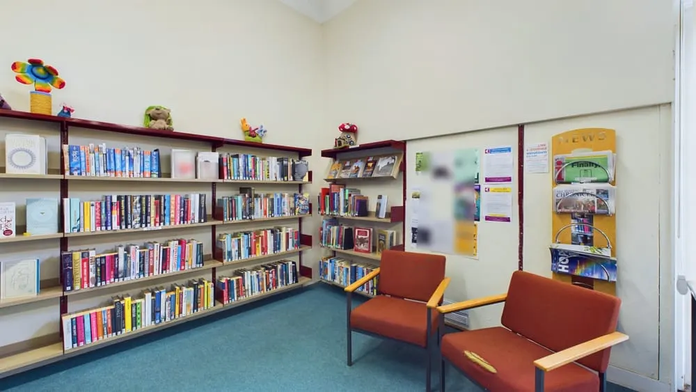 A library reading room with shelves of books, two rust-coloured armchairs, a turquoise floor, and colourful decorative items on the shelves.