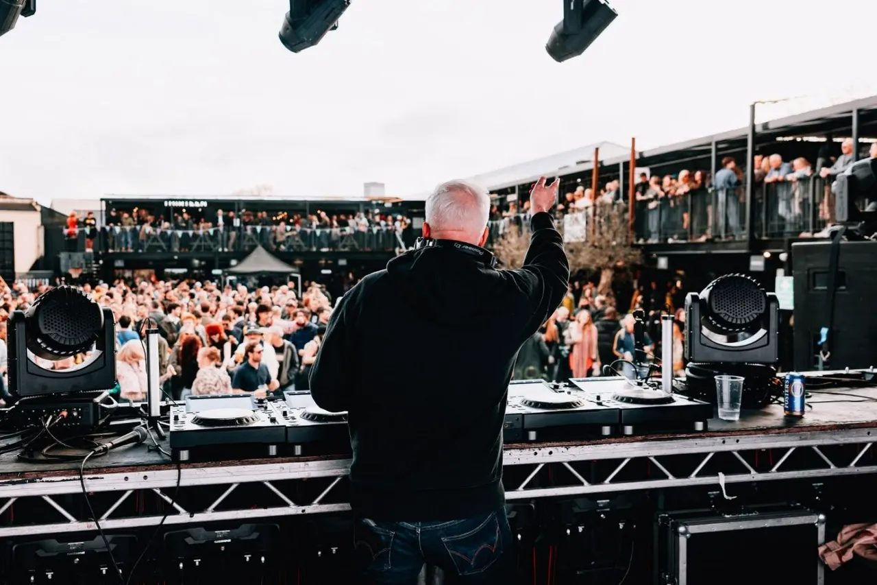 DJ with raised fist at decks on stage overlooking large crowd at outdoor event.