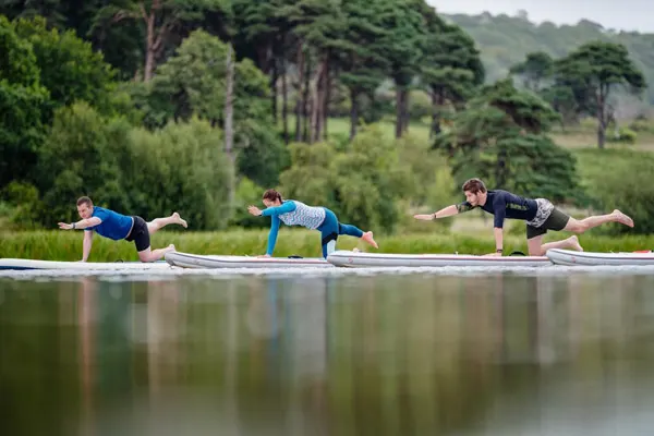 Three people practising yoga poses on a wooden platform beside a still lake, with forested hills behind.