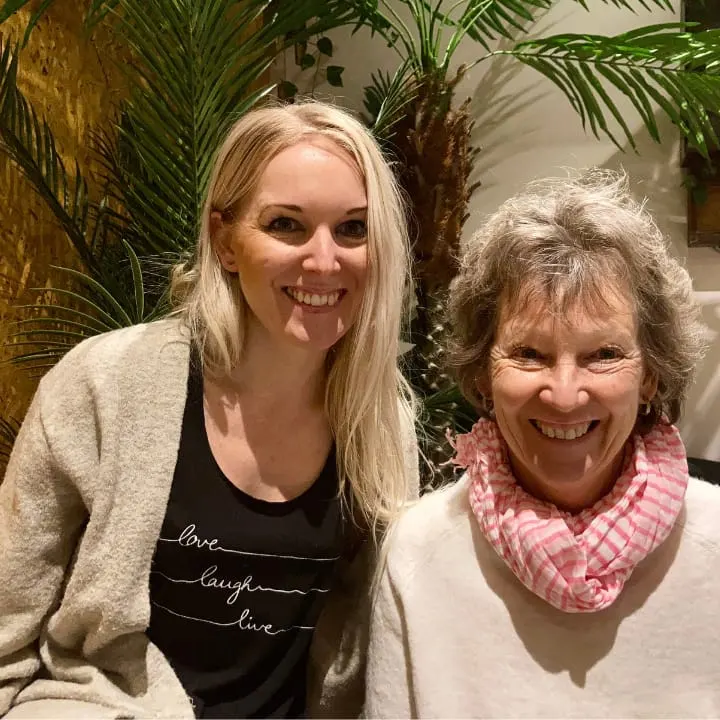 Two women smiling at the camera indoors with potted palm plants behind them.