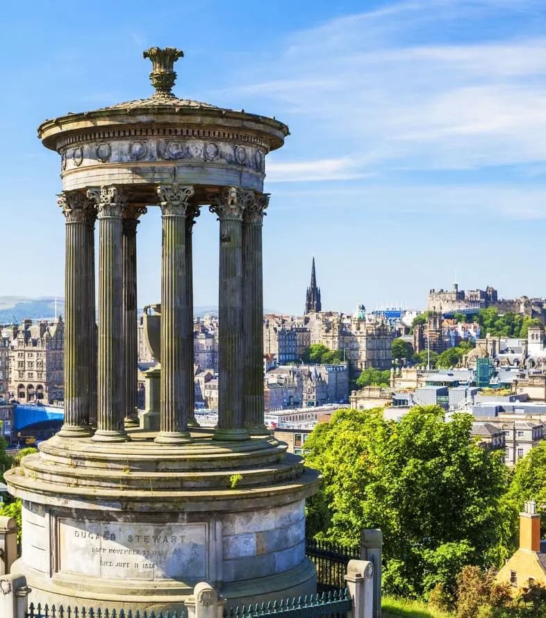Classical stone monument with fluted columns on a circular base, overlooking Edinburgh's cityscape with castle and church spires visible in the distance.