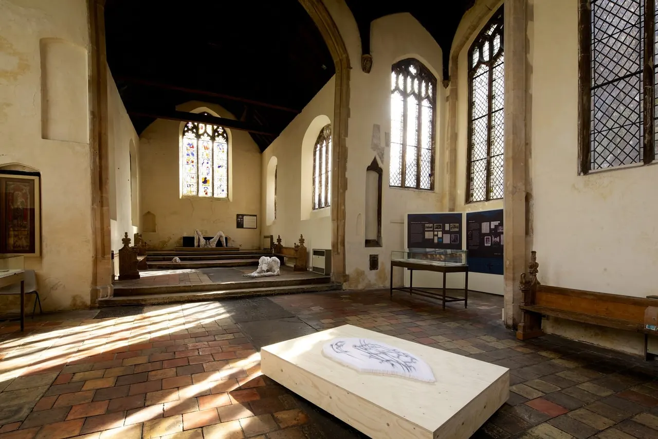 Interior of a medieval church with stone arches, stained glass windows, and display exhibits including artwork on a central plinth.