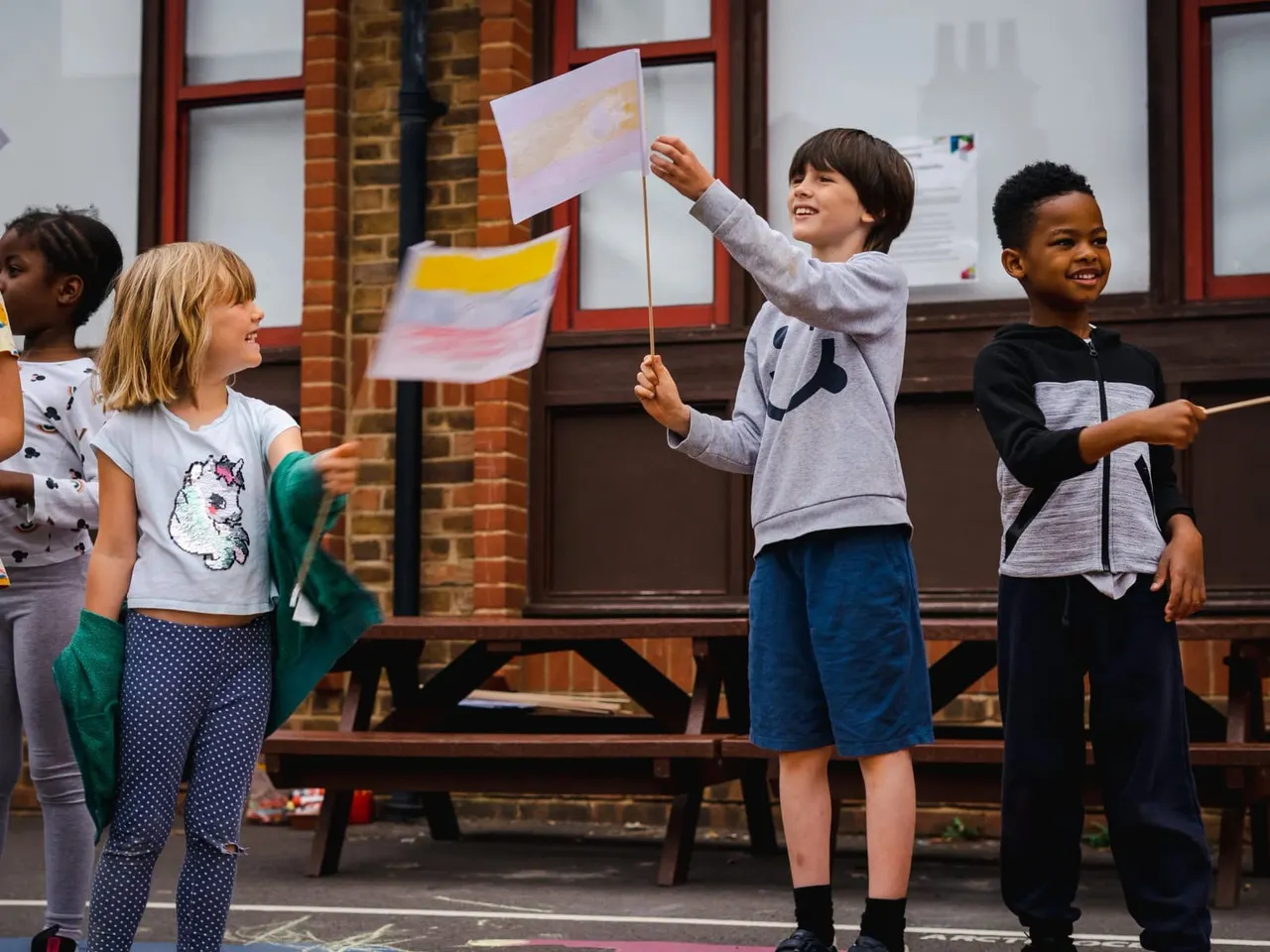 Four young children standing outdoors holding handmade flags with painted designs, smiling in front of a brick building.