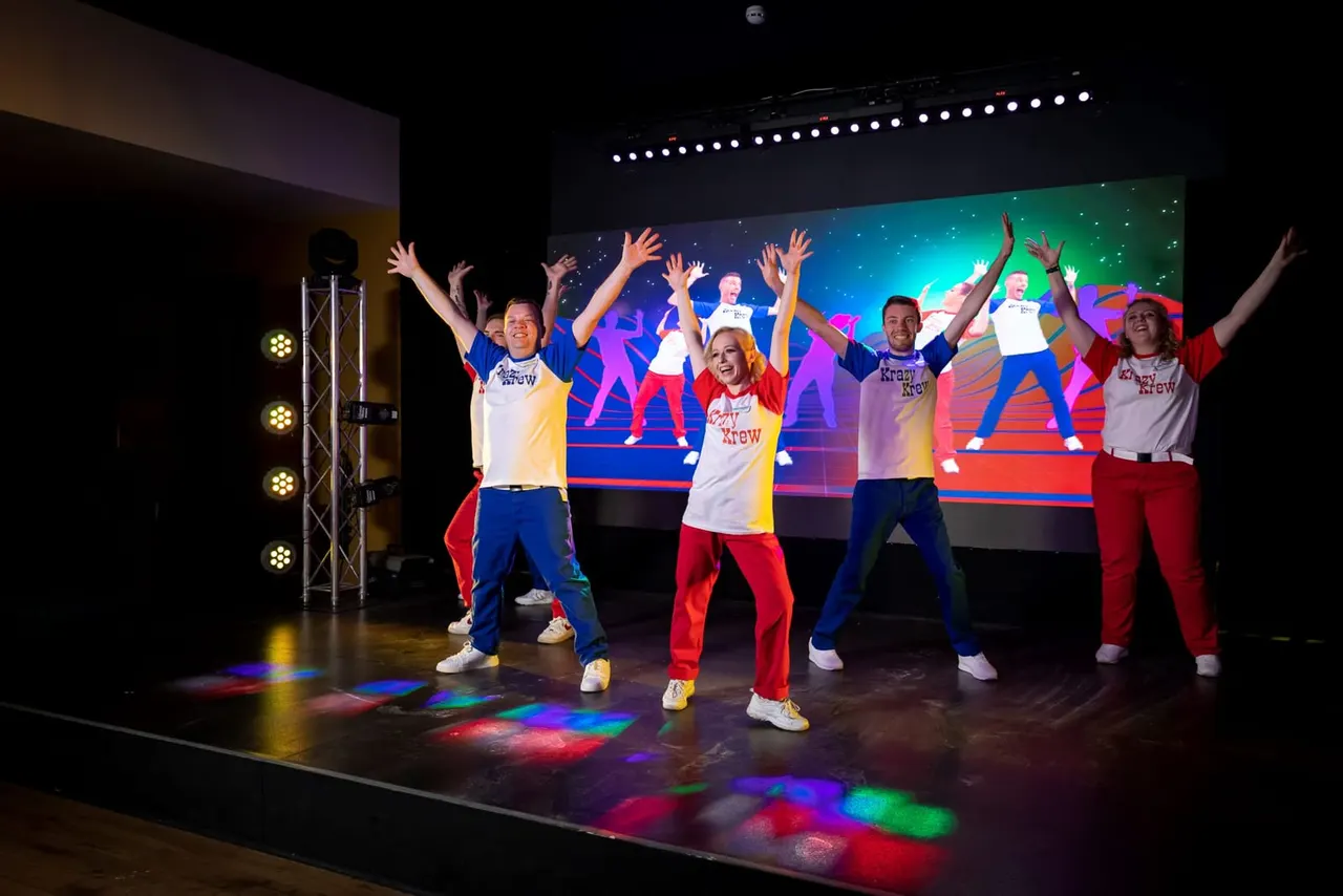 A group of performers dancing on stage with arms raised, wearing colourful matching outfits, with a large screen behind them and coloured stage lighting.